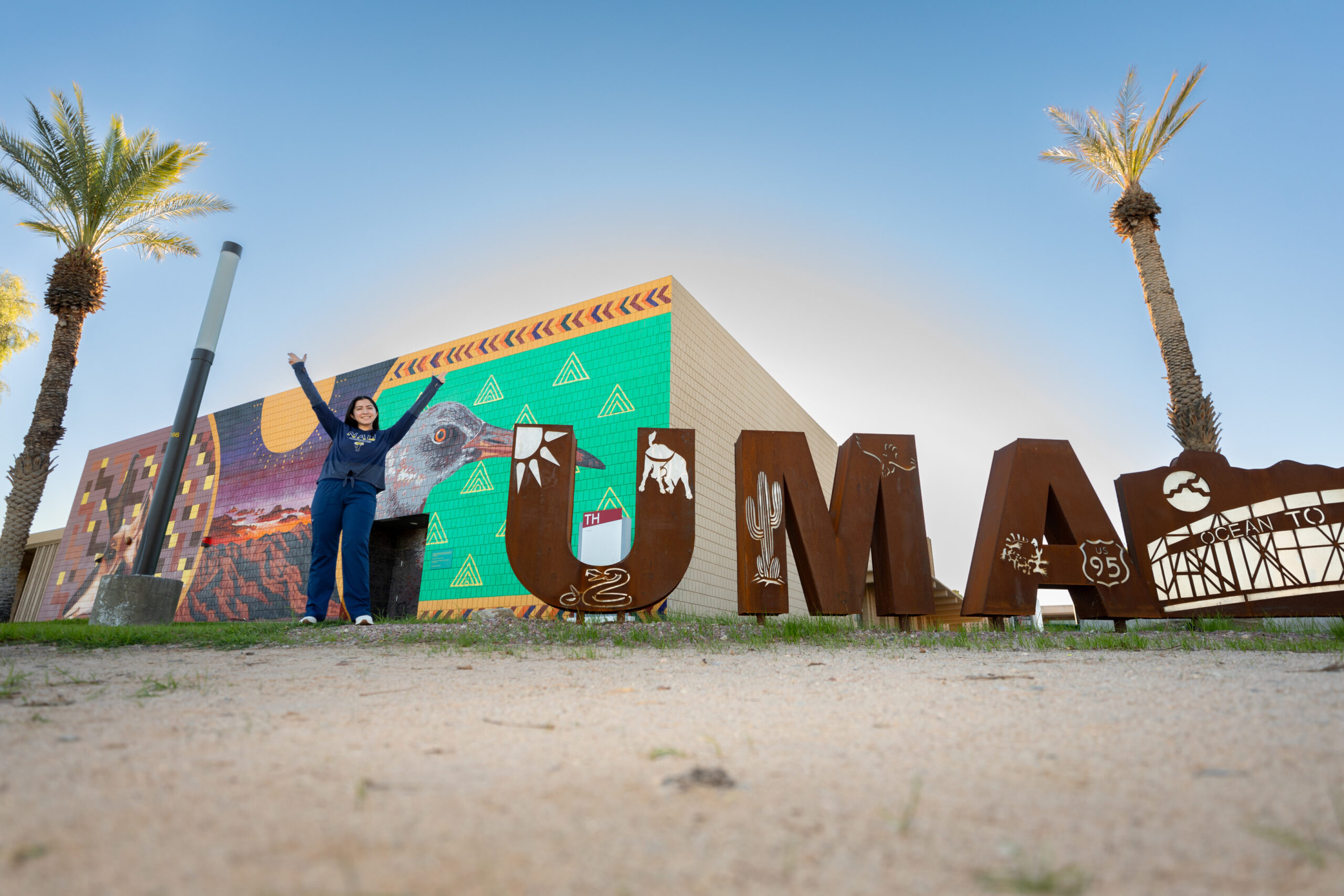 An NAU student makes a Y with her arms in the air next to a sign to spell out Yuma.
