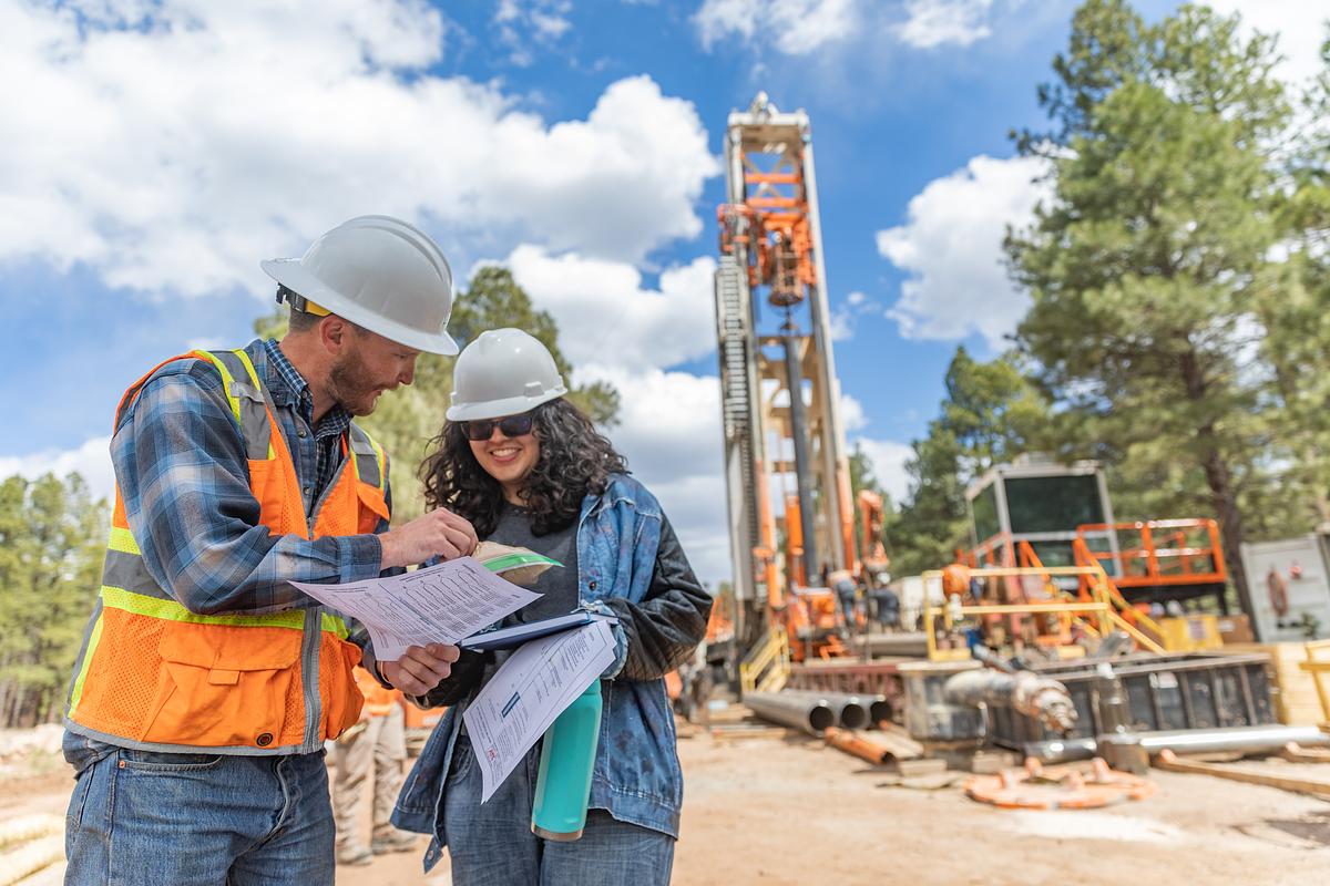 Construction workers wearing hardhats and high-vis vests review paperwork at a construction site.